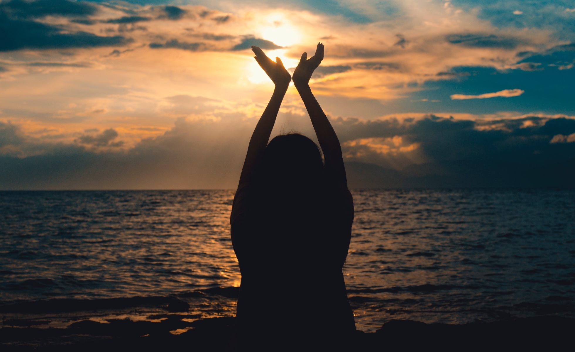 Silhouette of person against a sunset, holding up their hands to frame the setting sun