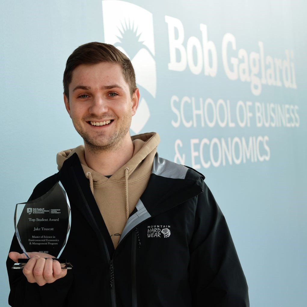 student stands in front of Bob Gaglardi School of Business and Economics sign and holds an award.