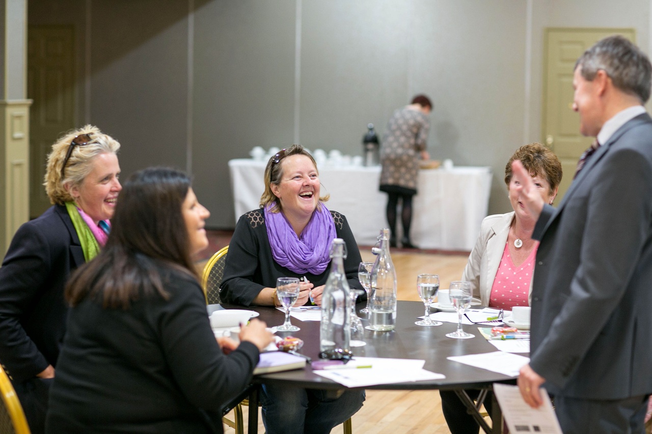A group of women sitting at a conference table, laughing as they chat with a man standing nearby.